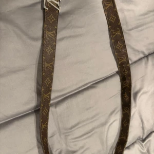 Brunt Louis Vuitton bälte i skinn - Säljer ett brunt Louis Vuitton bälte i skinn med klassiskt monogrammönster och silverfärgat spänne. Bältet har den ikoniska LV-loggan och är tillverkat i Frankrike. Perfekt accessoar för att lyfta din outfit. Säljs i befintligt skick, är använt men är fin utåt men lite sämre skick inåt alltså inget som syns. Priset är diskuterbart 