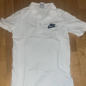 Vit pikétröja från Nike - Snygg vit pikétröja från Nike med klassisk krage och två knappar framtill. Tröjan har korta ärmar och en broderad Nike-logga i mörkblått på bröstet. Perfekt för en sportig och clean stil. Tillverkad i mjuk bomull för skön känsla.
