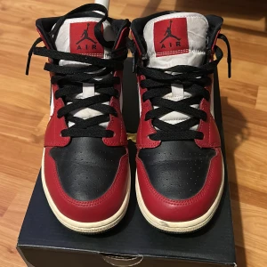Nike Air Jordan 1 mid (GS)  - Nike Air Jordan 1 Chicago i klassisk färgkombination med rött, svart och vitt skinn. Snygg svart swoosh, svart snörning och ikonisk Air Jordan-logga på sidan. Mids höjd och vit sula med svart yttersula. Perfekta för dig som vill sticka ut med en tidlös sneaker.