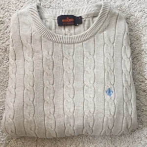 Beige kabelstickad tröja från Morris - Kabelstickad beige tröja från Morris i 100% pima bomull. Klassisk rund halsringning, ribbade muddar och diskret blå logga broderad på bröstet. Perfekt för dig som gillar stilren och tidlös design.