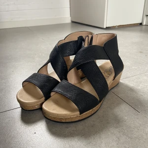 Svarta sandaler med korksula från Zoey - Snygga svarta sandaler från Zoey med breda korslagda remmar i syntetmaterial med lätt struktur. Skorna har öppen tå och en bekväm korksula med låg klack. Perfekta för varma dagar när du vill ha både stil och komfort.
