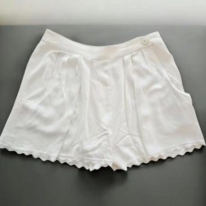 Vita shorts med spetskant - Superfina vita shorts från Guy Laroche med hög midja och söt spetskant längst ner. De har fickor på sidorna, veck framtill och en knappdetalj i midjan. Perfekta för varma sommardagar och ger en fräsch och luftig känsla. Materialet känns mjukt och lätt mot huden. Storlek 38