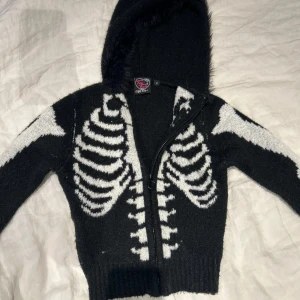 Svart kofta med skelettmönster från Too Fast - Svart zip up/hoodie/kofta med ett revbens skelettmönster i vitt med fluffig luva, knappt använd. Köpt för 750kr