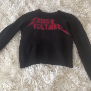 Svart tröja från Zadig & Voltaire - Snygg svart tröja från Zadig & Voltaire med röd text på framsidan. Perfekt för en lyxig  stil. Tröjan har långa ärmar och en klassisk rund halsringning.