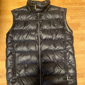 Svart dunväst från Parajumpers - Säljer en snygg svart dunväst från Parajumpers med dragkedja och hög krage. Västen har praktiska fickor med dragkedjor och en stilren design. Perfekt för kyliga dagar när du vill hålla värmen utan att kompromissa med stilen.