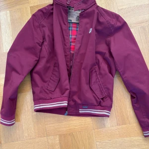 Vinröd bomberjacka med rutigt foder från Lambretta - Snygg vinröd bomberjacka med ett klassiskt rutigt foder i rött, blått och vitt. Jackan har dragkedja framtill och ribbade muddar vid ärmslut och nederkant. Perfekt för en stilren look!