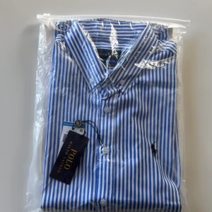 Blåvit randig skjorta från Ralph Lauren stl M - Snygg blåvit randig skjorta från Ralph Lauren med klassisk krage och knappar framtill. Perfekt för en stilren look. Den har en broderad logga på bröstet. Stl M classic fit.