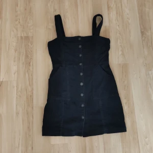 Svart bodyconklänning - Säljer en snygg svart manchesterklänning från Divided HM med knappar framtill. Klänningen har breda axelband och en figurnära passform. Perfekt för en stilren look!