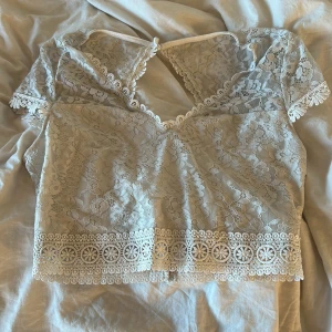Vit spetsblus - Säljer en vacker vit spetsblus med korta ärmar och fina spetsdetaljer. Blusen har en v-ringad design och en dekorativ kant nedtill. Perfekt för en elegant look.