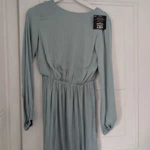 Ljusblå klänning från N.Y. Trend - Säljer en elegant ljusblå/grön klänning från N.Y. Trend med långärmad design och v-ringad rygg. Klänningen har en elastisk midja för en bekväm passform. Perfekt för en stilren look. Helt ny med prislappen kvar 
