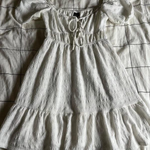 Vit klänning från Gina Tricot - Charmig vit klänning från Gina Tricot med korta ärmar. Klänningen har en söt volangdetalj vid fållen och knytning framtill för en romantisk look. Perfekt för varma sommardagar! Väldigt fint skick och sällan använd.