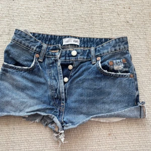 Blå jeansshorts från H&M - Säljer ett par blå jeansshorts från Zara  med fransiga kanter och knappar framtill. Shortsen har en klassisk femficksdesign och är perfekta för sommardagar. De har en avslappnad och trendig look.