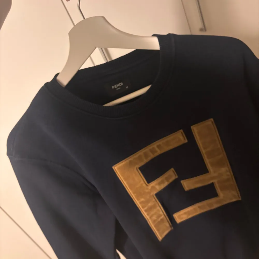 Säljer en snygg mörkblå tröja från Fendi med ett stort guldigt Fendi-logotyp på framsidan. Tröjan har långa ärmar och en klassisk rund halsringning. Perfekt för en stilren look.. Neuletakit & Villapaidat.