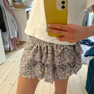 Blommig kortkjol med volang - Söt blommig kortkjol i lila, vit och gul med volangdetaljer. Kjolen har en elastisk midja för en bekväm passform. Perfekt för varma dagar och ger en romantisk touch till din outfit.