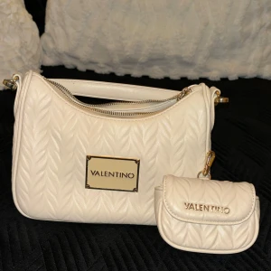 Vit quiltad axelväska från Valentino - Snygg vit/beige axelväska från Valentino med quiltad design och gulddetaljer. Väskan har en praktisk dragkedja och en justerbar axelrem. Perfekt för att ge din outfit en lyxig touch. Aldrig använt.
