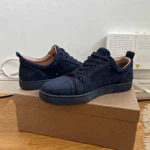 Mörkblå sneakers med röd sula - Snygga mörkblå sneakers i mocka med en ikonisk röd sula. Skorna har snörning och en rund tå, vilket ger en klassisk look. Perfekta för dig som vill ha stil och komfort i ett.