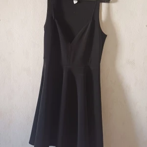 Svart klänning från NLY One - Snygg svart klänning från NLY One med en v-ringad design och ärmlös stil. Perfekt för en elegant look. Klänningen har en kort längd och en skön passform. Obs sydd i axlarna 