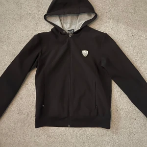 Svart hoodie från Emporio Armani - Snygg svart hoodie från Emporio Armani med dragkedja och huva. Den har ett stilrent märke på bröstet och är perfekt för en casual look. Insidan av huvan är grå vilket ger en fin kontrast.
