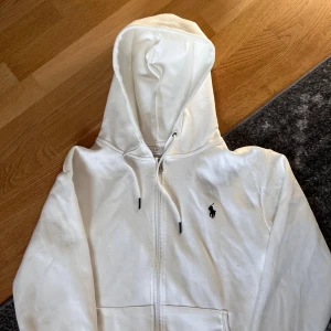 Vit hoodie från Ralph Lauren - Säljer en stilren vit hoodie från Ralph Lauren med dragkedja och det klassiska logotypen broderad på bröstet. Perfekt för en avslappnad look. Hör av dig vid intresse!