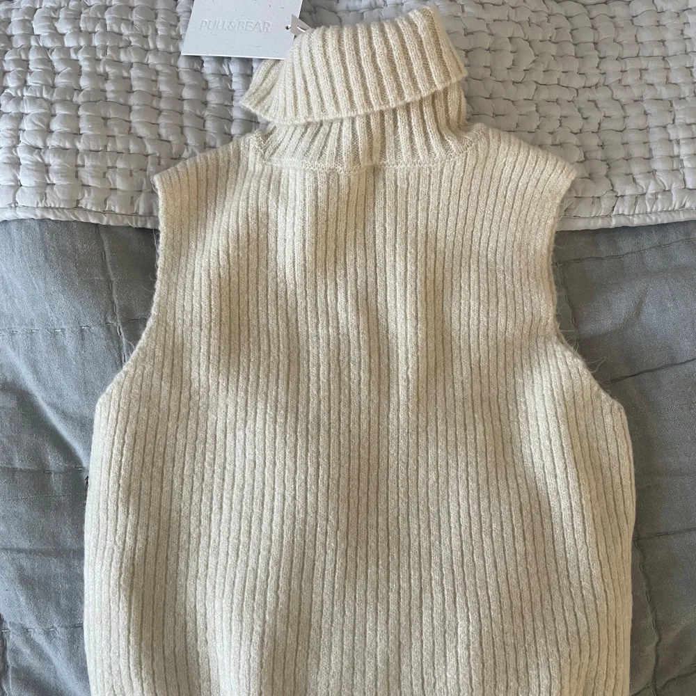 Snygg beige stickad väst med ribbad design och polokrage. Perfekt för lager-på-lager. Västen är ärmlös och har en mjuk och bekväm passform.. Neuleet.