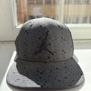 Grå keps med stänkmönster från Jumpman - Snygg grå keps med svart stänkmönster och Jumpman-logga framtill. Justerbar passform med spänne bak. Perfekt för en sportig look eller casual stil.