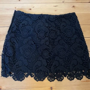 Svart spetskjol - Snygg svart kortkjol i spets med vackert mönster. Perfekt för att ge en elegant touch till din outfit. Kjolen har en fin kantdetalj längst ner som ger extra stilpoäng. 