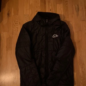 Svart quiltad jacka från Nike - Säljer en svart quiltad jacka från Nike med dragkedja och huva. Jackan har ett stilrent Nike-märke på bröstet och är perfekt för kyligare dagar. Den är långärmad och tillverkad i syntetmaterial.