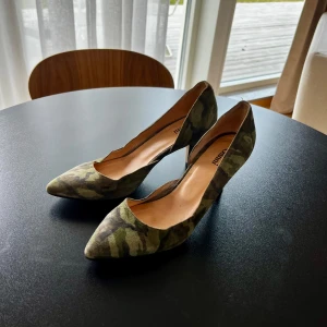 Camouflage pumps från Ganni - Snygga pumps från Ganni i ett coolt camouflage-mönster. Skorna har en spetsig tå och en elegant klack, perfekt för att ge din outfit en unik touch. De är tillverkade i ett mjukt material som ger en bekväm passform. Perfekta så som till ett par jeans eller utgång! Nypris ca 2000