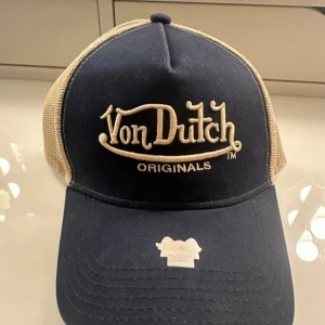 Blå och beige truckerkeps från Von Dutch - Snygg truckerkeps från Von Dutch i blått och beige. Kepsen har ett klassiskt mesh-tyg baktill och en broderad logga framtill. Perfekt för en avslappnad stil.