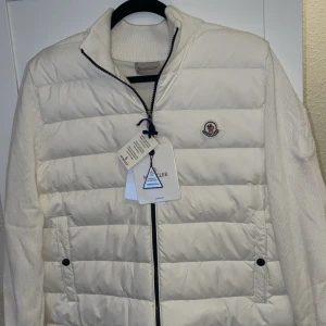 Moncler cardigan vit - Snygg vit dunväst från Moncler med dragkedja och stickade ärmar. Västen har en broderad logotyp på bröstet och är perfekt för kyligare dagar. Den har en stilren design med ribbade detaljer vid ärmslut och nederkant.