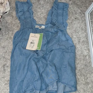 Blå ärmlös blus från H&M - Säljer en söt blå ärmlös blus från H&M med volangdetaljer på axelbanden. Perfekt för varma sommardagar. Blusen har en luftig passform och är tillverkad i ett lätt material. Ny och har fortfarande prislappen kvar (kostade från början 349).