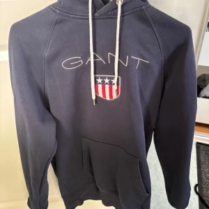 Mörkblå hoodie från GANT - Säljer en snygg mörkblå hoodie från GANT med en stor logga på framsidan. Hoodien har en praktisk magficka och justerbar huva med vita snören. Perfekt för en avslappnad stil.