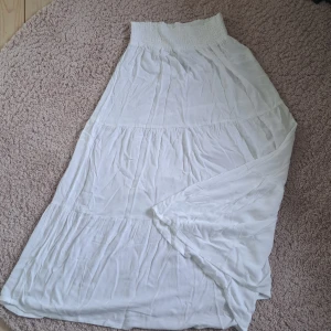 Vit långkjol från Lager 157 - Säljer en vit långkjol från Lager 157, modell Bella. Kjolen har en elastisk midja och är tillverkad i ett lätt och luftigt material, perfekt för varma dagar. Den har en enkel och stilren design som passar till många olika tillfällen.