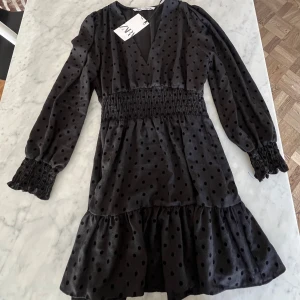 Svart prickig klänning från Zara - Säljer en kort svart klänning från Zara med prickigt mönster och v-ringning. Klänningen har långa ärmar och volangdetaljer vid fållen. Den elastiska midjan ger en smickrande passform. Perfekt för en stilren look! Köpt för 400, aldrig använd!!! 