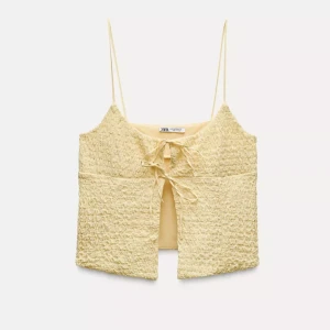 Gul topp med knyt från Zara - Säljer en söt gul topp från Zara med smala axelband och knytning framtill. Toppen har en texturerad yta. Storlek L, men passar m.