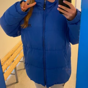 Blå pufferjacka från Helly Hansen - Säljer en blå pufferjacka från Helly Hansen med dragkedja framtill. Jackan har en hög krage och långa ärmar, perfekt för kyliga dagar. Den är både stilren och funktionell.