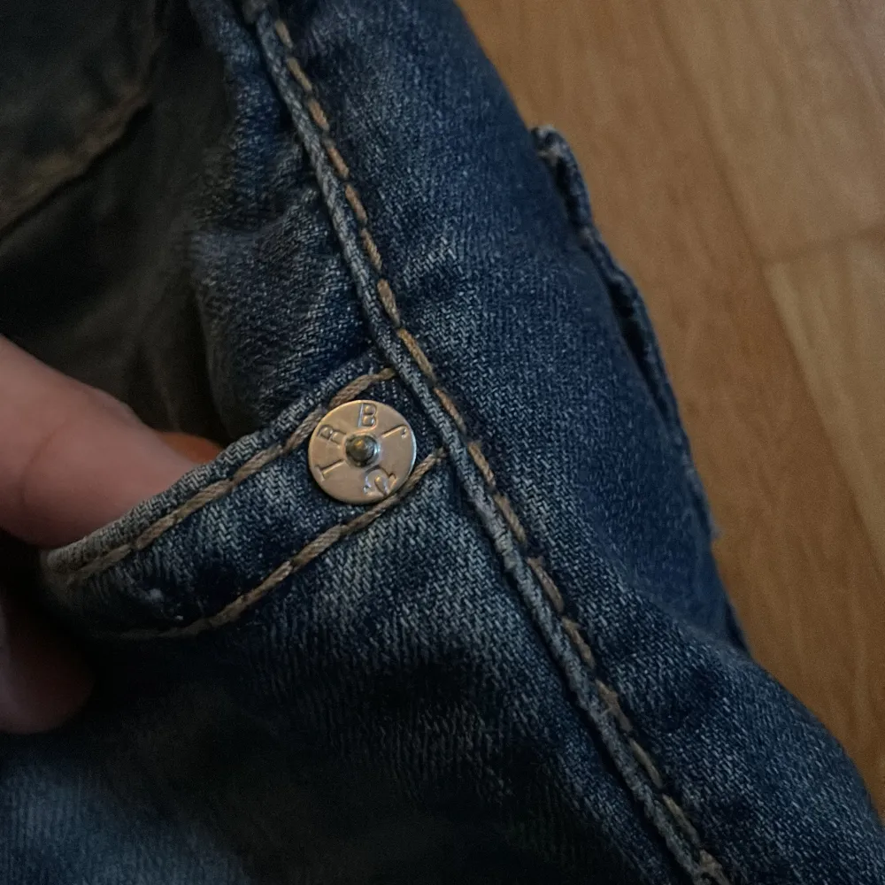 Säljer ett par snygga blå jeans från True Religion med klassisk femficksdesign och knappar. Jeansen har en straight passform och är tillverkade i slitstarkt denim. Perfekta för en avslappnad stil.. Farkut & Housut.