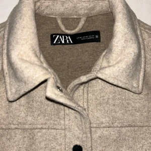 Beige jacka från Zara - Felfri, knappt använd beige jacka från Zara i en stilren design. Jackan har en klassisk krage och knappar framtill. Två stora fickor med lock på framsidan ger en cool look. Perfekt för våren som kommer nu!
