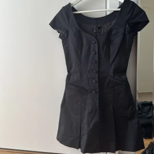 Svart klänning från Tiger of Sweden - Elegant svart klänning från Tiger of Sweden med knappar framtill och korta ärmar. Perfekt för en stilren look. Klänningen har en klassisk design med en lätt utställd kjol.