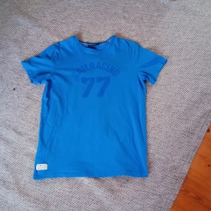 Blå t-shirt från Sail Racing - Säljer en blå t-shirt från Sail Racing med tryck 'SAIL RACING 77' på framsidan. T-shirten är kortärmad och har en rund halsringning. Perfekt för en avslappnad stil.