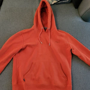 Orange Superdry hoodie storlek L - Säljer en snygg orange hoodie från Superdry med broderad logga på bröstet. Den har en klassisk känguruficka och justerbar huva med snören. Perfekt för en avslappnad stil.