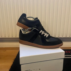Svarta sneakers från Maison Martin Margiela - Snygga svarta sneakers från Maison Martin Margiela med vita snören och en brun sula. Skorna har en stilren design och är perfekta för en trendig look. Levereras med originalförpackning och skyddspåsar.
