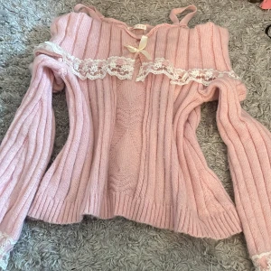 Rosa stickad offshoulder-tröja med spetsdetaljer - Säljer en söt rosa stickad offshoulder-tröja med spetsdetaljer och en rosett framtill. Tröjan har långa ärmar och ribbad struktur, perfekt för en romantisk look. Passar bra till både jeans och kjol. 💖