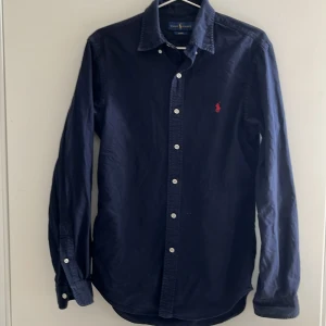 skjorta Ralph Lauren - Säljer en stilren mörkblå skjorta från Ralph Lauren med klassisk krage och knappar framtill. Skjortan har en broderad röd logotyp på bröstet och är i slim fit. Perfekt för en elegant look.
