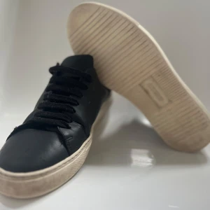 Svarta sneakers från Axel Arigato - Snygga svarta sneakers från Axel Arigato med en stilren design. Yttersulan är vit och ger en snygg kontrast till det svarta skinnet. Perfekta för en casual look. Skorna har några defekter. Hör av dig vid minsta fundering, priset är ej hugget i sten! 😉