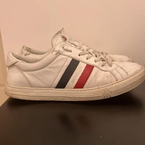 Moncler skor - Snygga vita sneakers från Moncler med klassiska ränder i blått och rött på sidan. Skorna har snörning och rund tå, vilket ger en stilren look. Perfekta för en avslappnad men ändå stilfull stil. Nypris på dessa var 3900kr men säljs för 550kr pga att de är slitna, en bit av sulan är av men de fungerar fullt och läcker inte in vatten, 6/10 skick