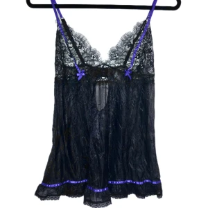 Svart spetslinne med lila detaljer - Elegant svart spetslinne med lila axelband och rosetter. Det har en vacker spetsdesign och en liten volangkant nedtill. Perfekt för en speciell kväll. Helt oanvänd bara testad på. Man kan knäppa i ryggen som på en bh.