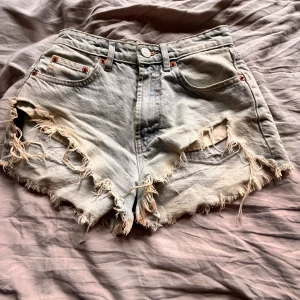 Ljusa jeansshorts med fransar - Säljer ett par ljusa jeansshorts med fransiga kanter och slitna detaljer. Shortsen har en knapp och dragkedja framtill samt klassiska fem fickor. Perfekta för en avslappnad sommarlook!