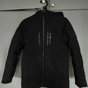 Svart dunjacka från Sail Racing - Säljer en svart dunjacka från Sail Racing med huva och två bröstfickor med dragkedja. Jackan har Sail Racing-logga på bröstet och ett Sail Antarctica-märke på ärmen. Fodrad med 70% dun och 30% fjäder, yttermaterial i polyester och spandex. Perfekt för kalla dagar.