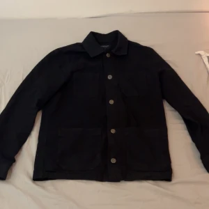 Svart overshirt med knappar - Stilren svart overshirt med klassisk krage och fyra fickor framtill. Jackan har raka linjer, knäppning med metallknappar och lång ärm. Perfekt för lager-på-lager och enkel att matcha med olika outfits.